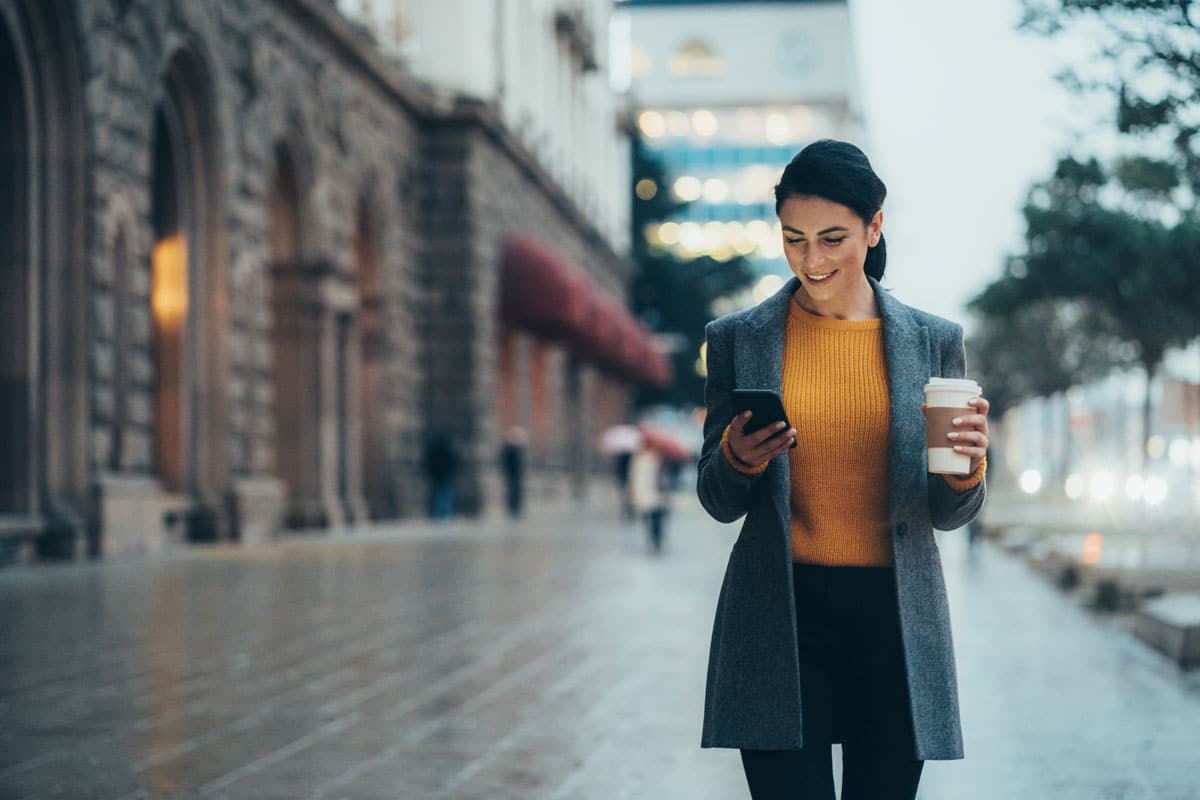 happy woman walking down the street checking her phone and holding a cup of coffee